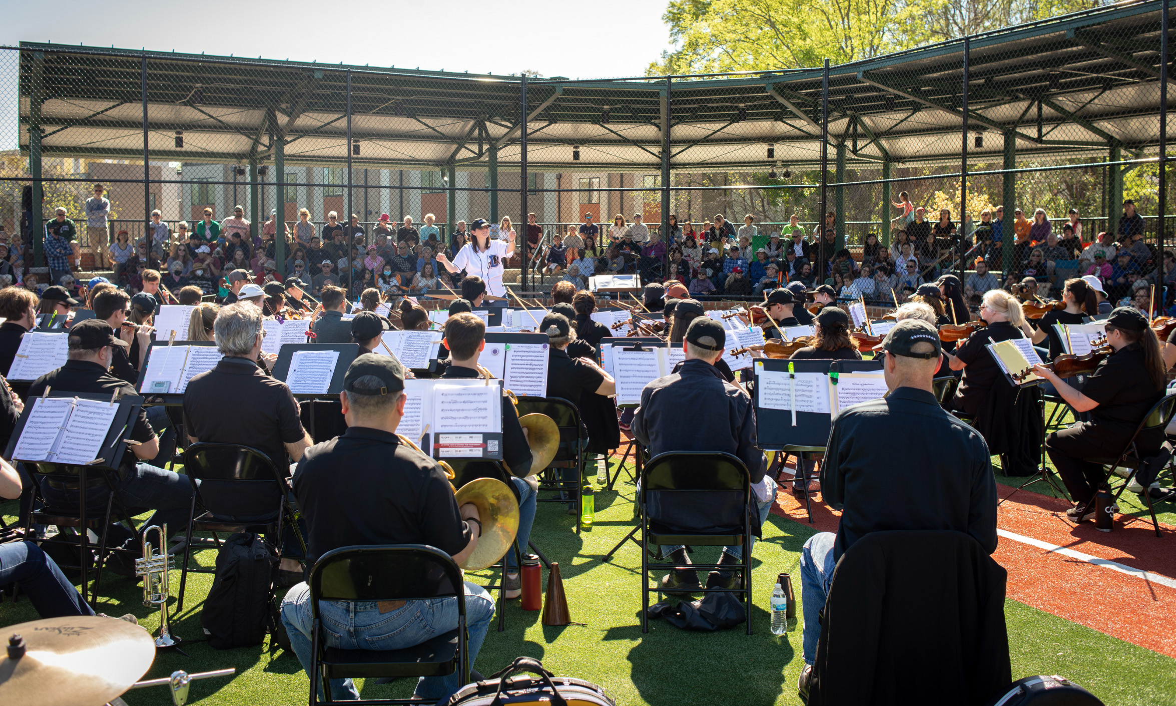 An orchestra performs outdoors at a small baseball stadium