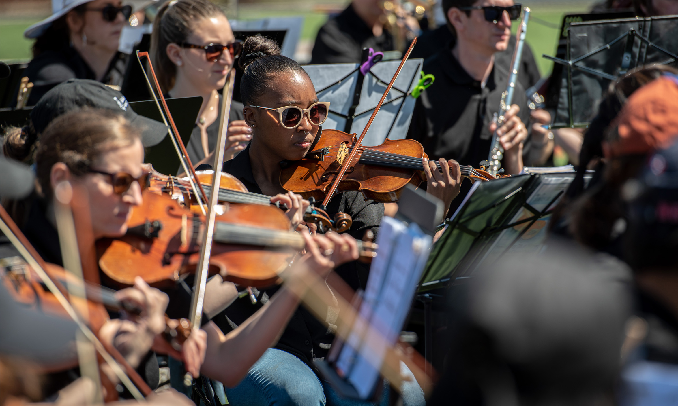 An orchestra plays outdoors as members wear sunglasses.