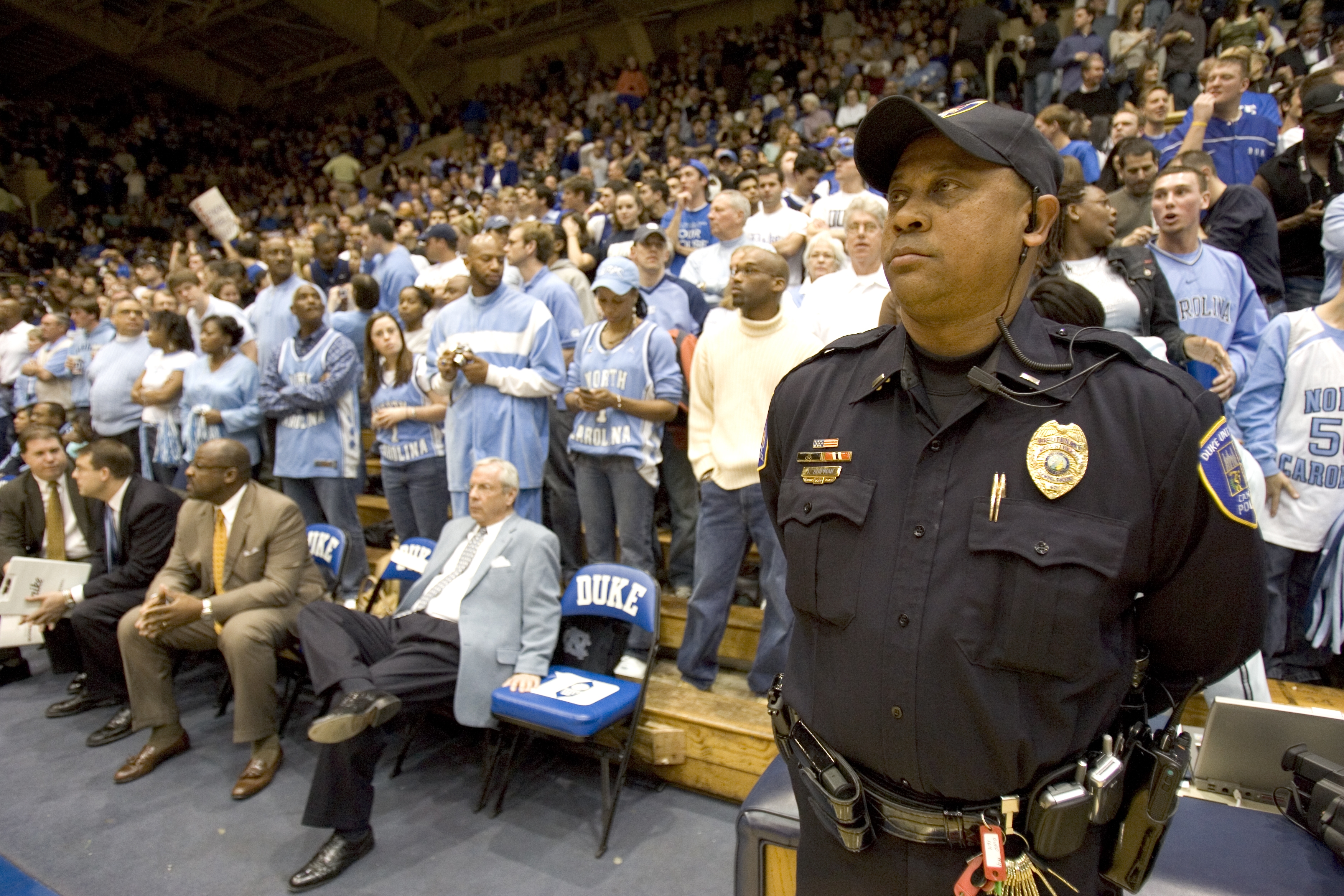 A police officer stands on the edge of a group of North Carolina fans at a basketball arena.