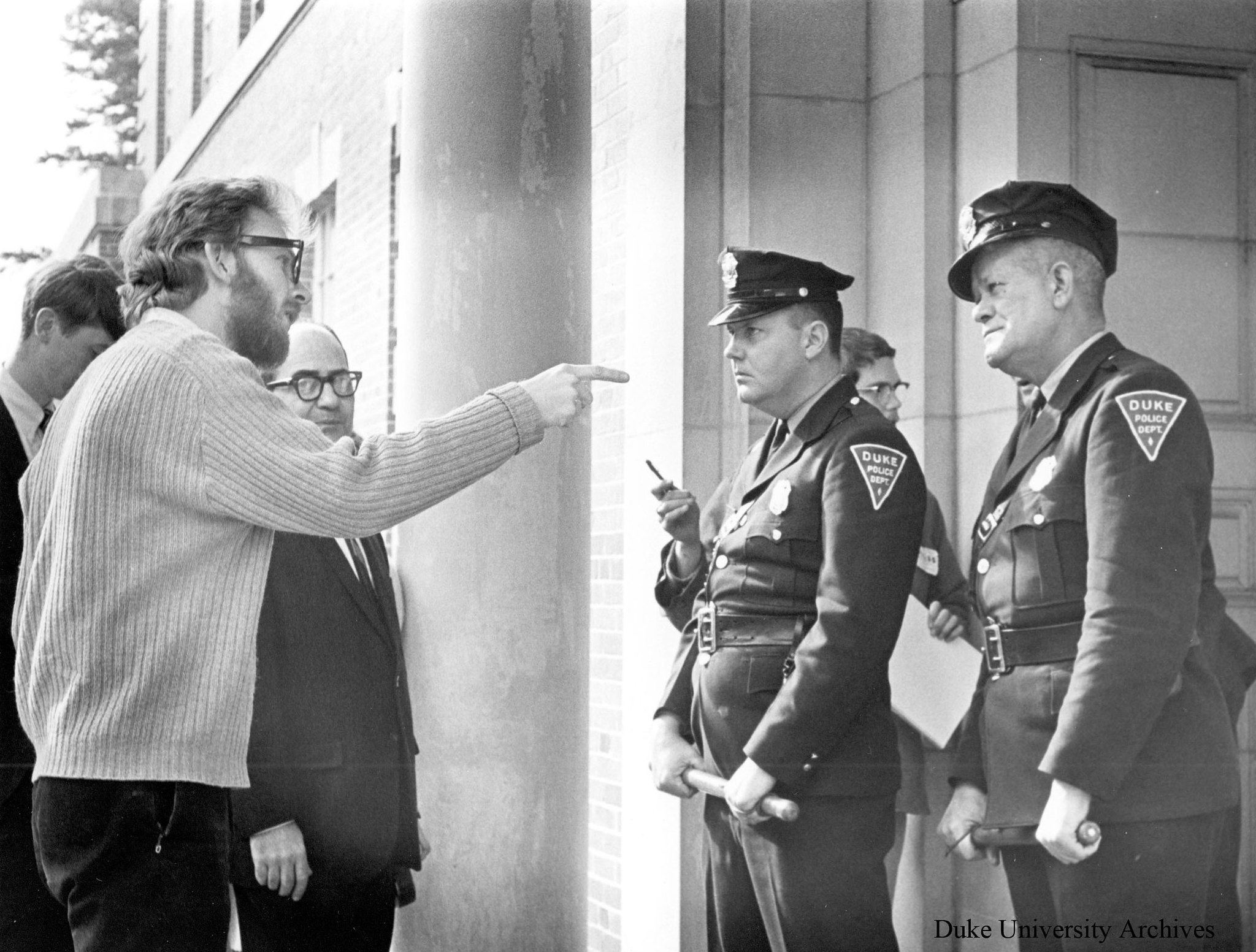 A man wearing glasses points at two Duke Police officers standing at the entrance to a building.
