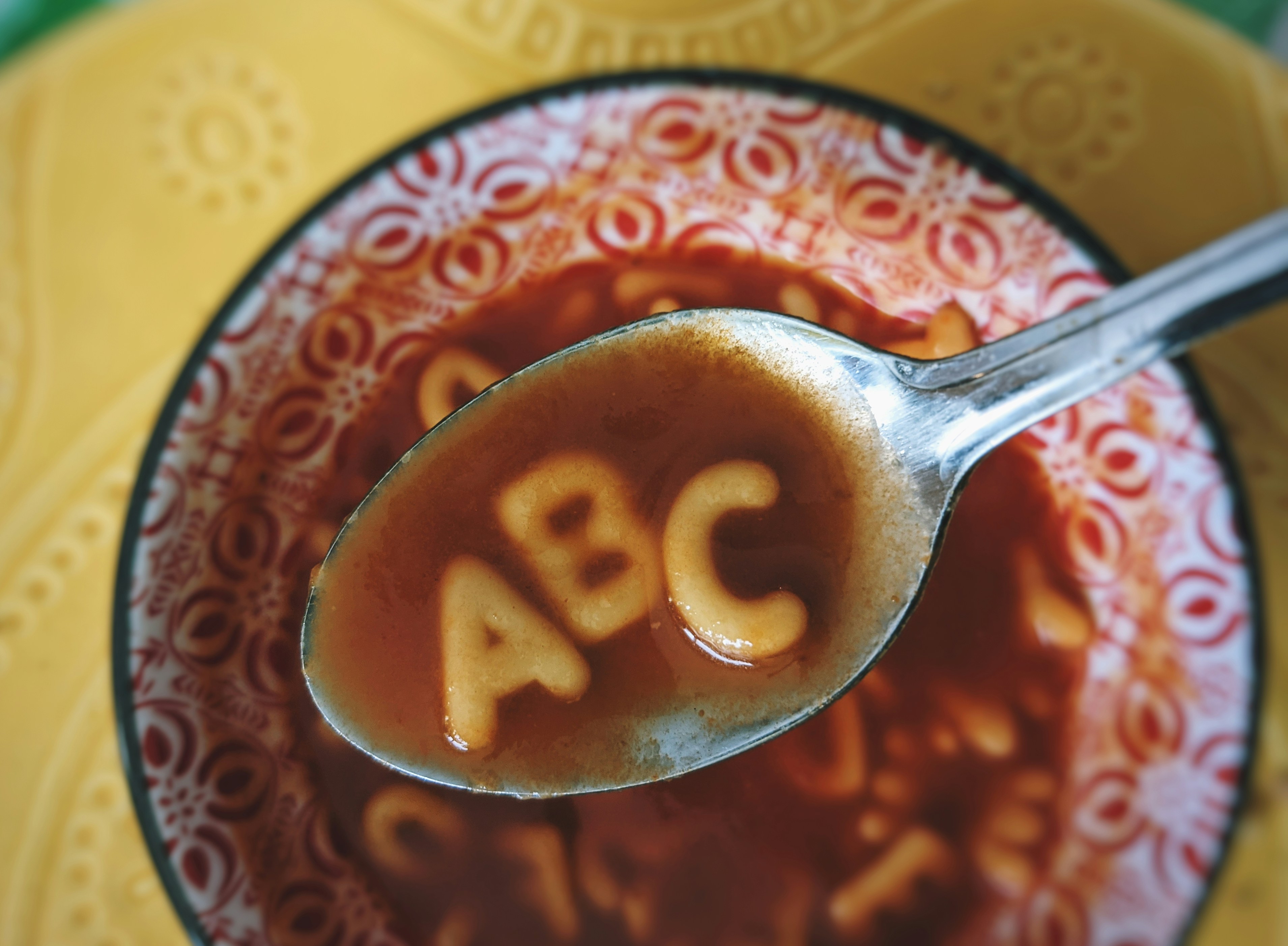 A bowl of soup with a spoon holding noodles shaped as A, B and C.