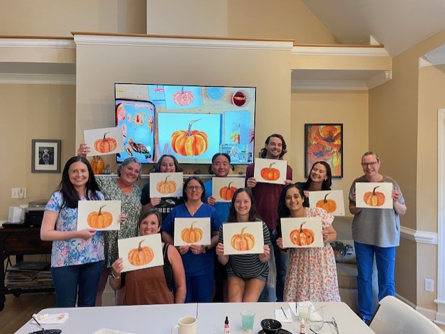 A group of people hold paintings of a pumpkin while a TV displaying the same pumpkin is behind them.