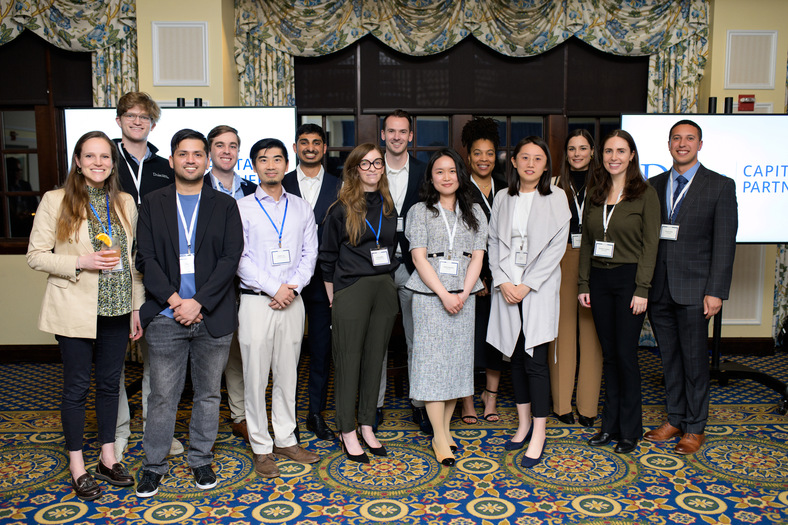 Fourteen people stand in professional attire in two rows, looking at the camera smiling. They are at the Washington Duke Inn, with ornate carpeting and two screens behind them with the Duke Capital Partners logo in Duke blue on white.