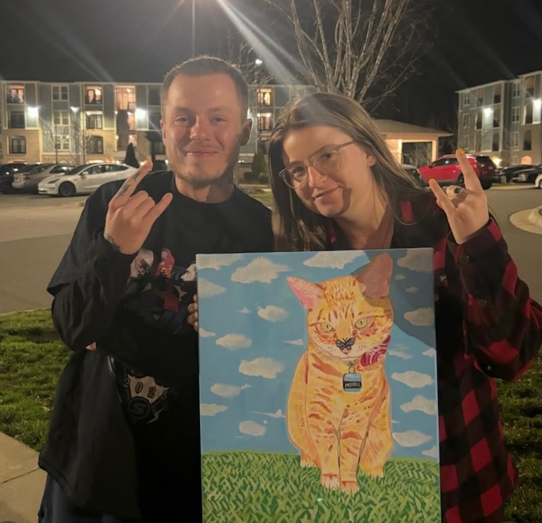 A man and a woman stand as the woman holds a painting of an orange tabby cat