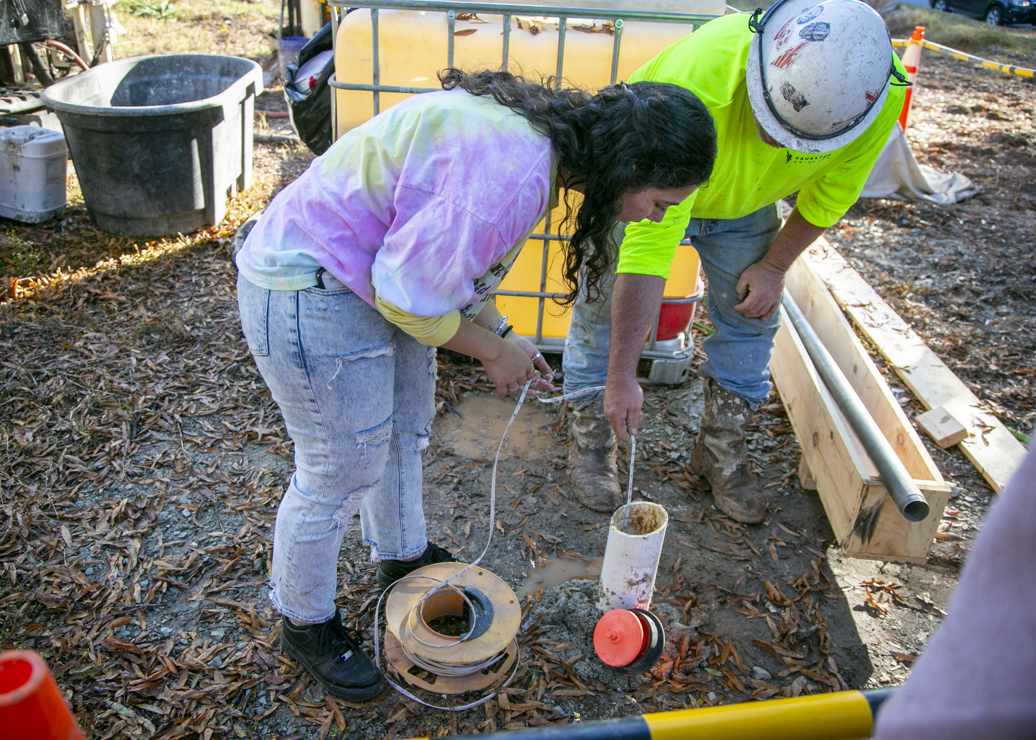 A student leans over the pvc pipe coming out of the ground and lowers a measuring tape down the hole while a hardhat wearing engineer guides.
The ground is wet and muddy and drilling material is all around them.