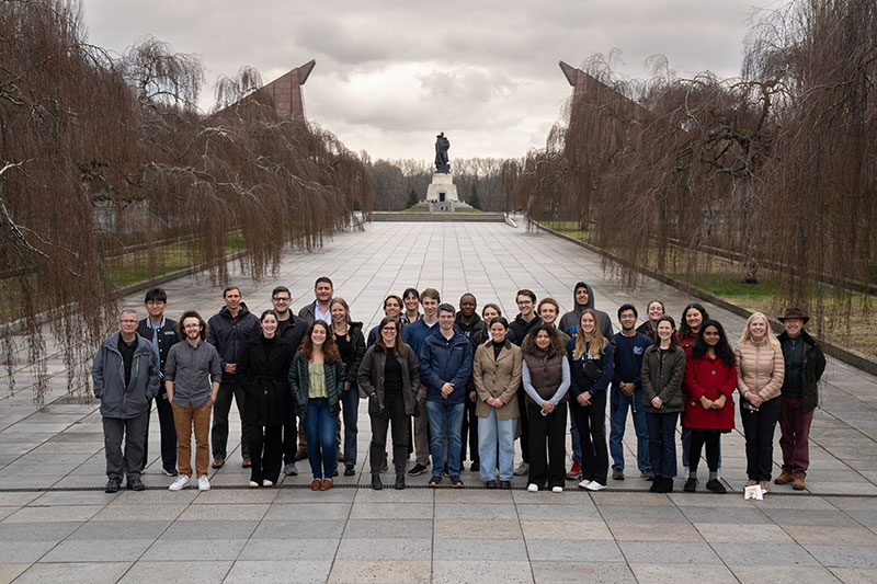 A group of students and other members of the trip stand in a group on a wide stone walkway surrounded by bare trees with a statue in the distance nin the background