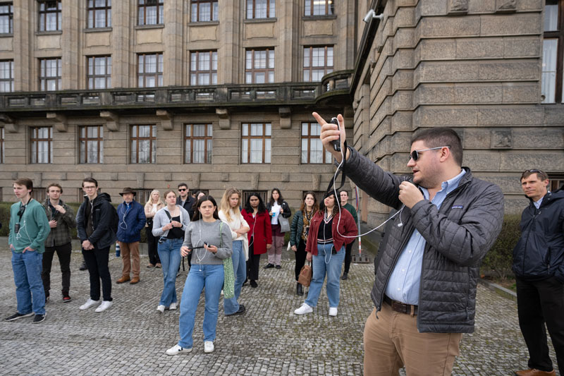 A man stands on a cobble stone street talking and pointing to the right with a group of students in the background