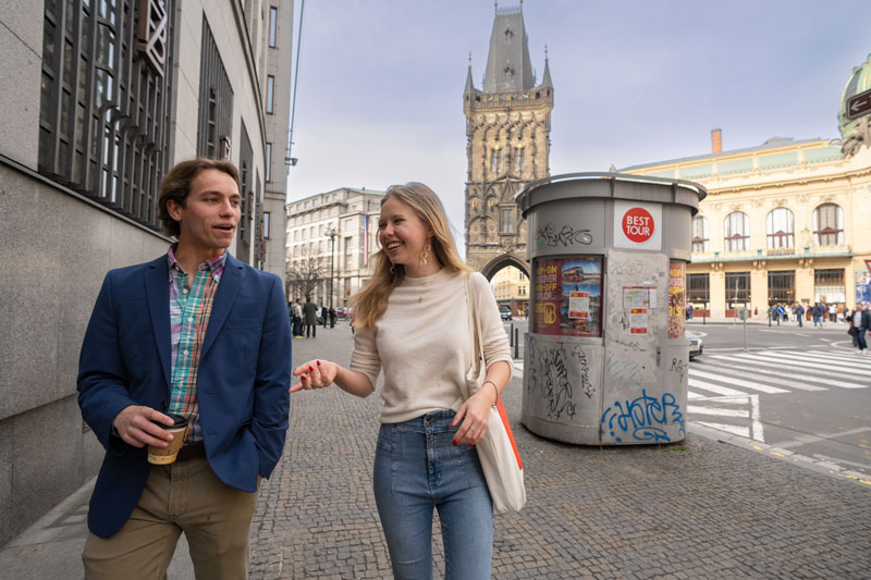Two students casually walking down the cobbled streets of Prague with a 550 year old tower in the background