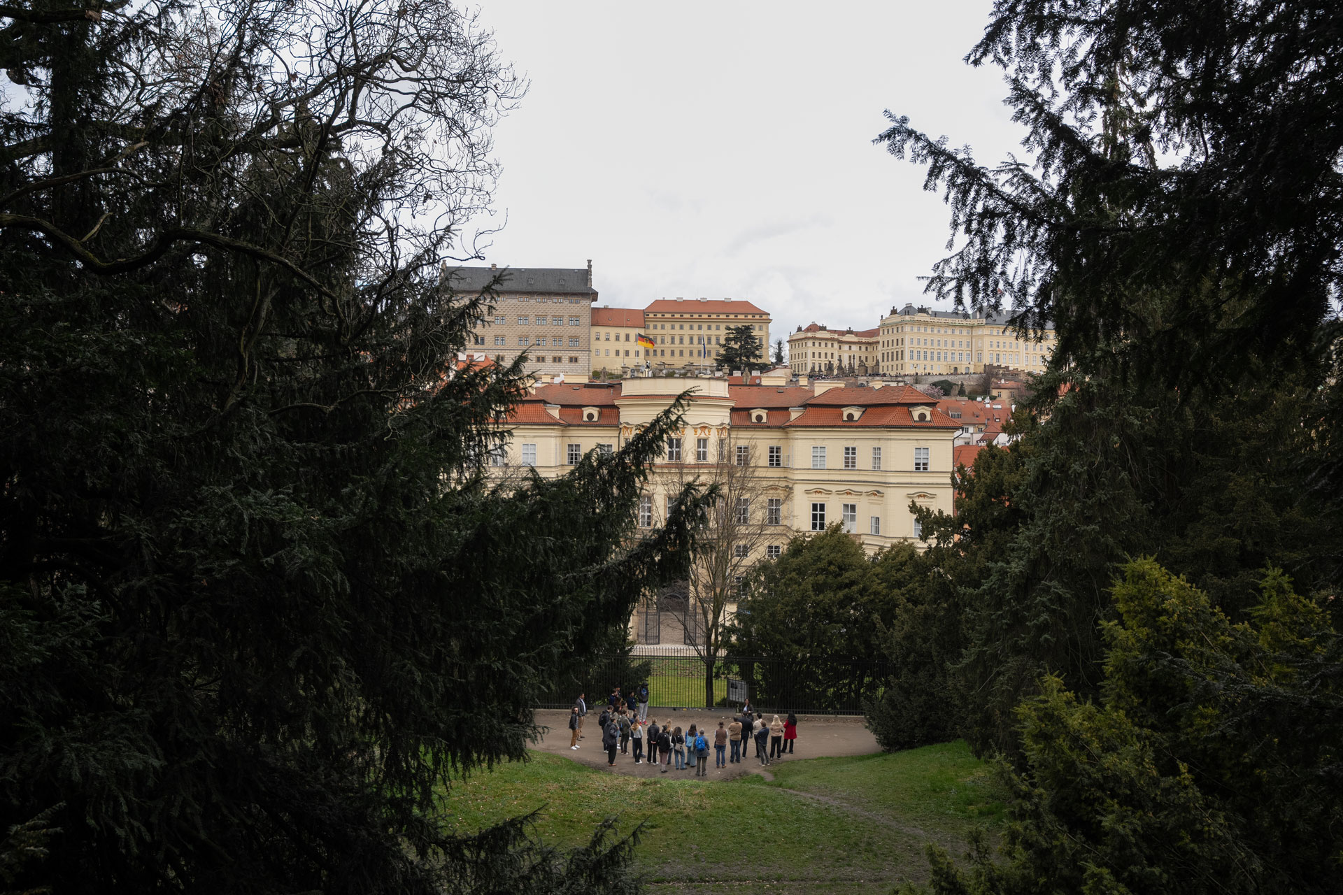 The group stands at the bottom of the hill in front of the gate for the Embassy of Germany. Large evergreen trees flank the sides of the photo.
