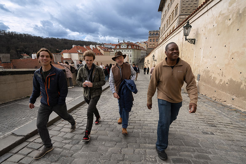 Four members of the staff ride stride up the cobble stone street under a cloudy sky