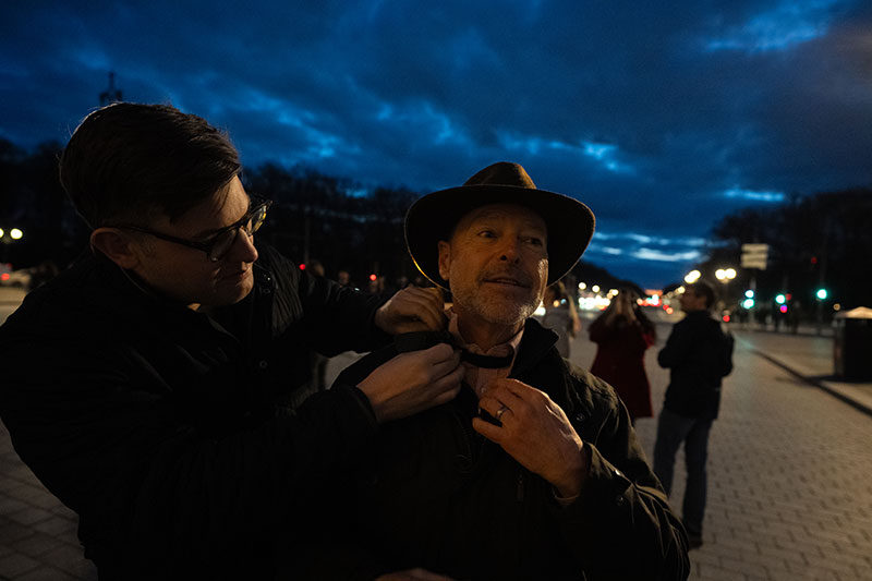One person is using two hands to tie a bow tie around another persons neck on a city street at dusk