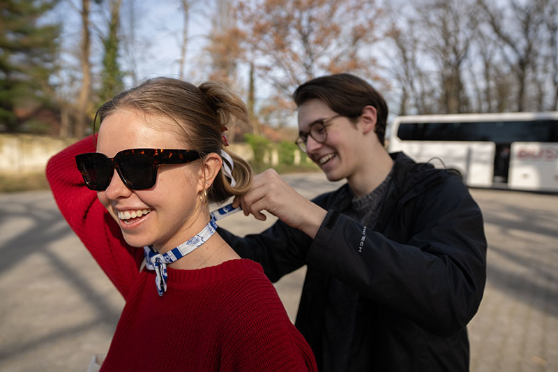 One person stands behind another securing a bow tie on a street in daylight