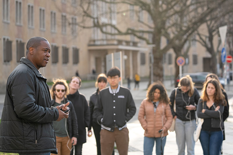 A ride member stands in the foreground with students out of focus in the background listening