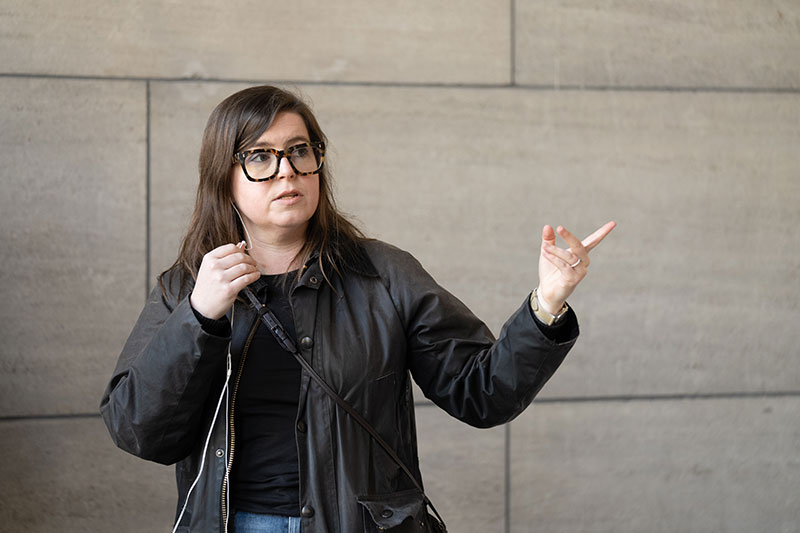 A woman in a dark jacket stands in front of a stone wall pointing to the right