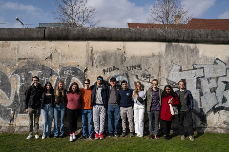 A group of students poses lined up, posing for a photo in front of the graffitied Berlin Wall