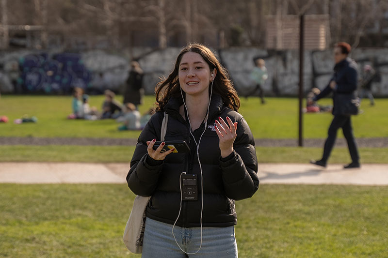 A student stands in a grassy area speaking to the group as people enjoy the green space with the Berlin Wall in the background