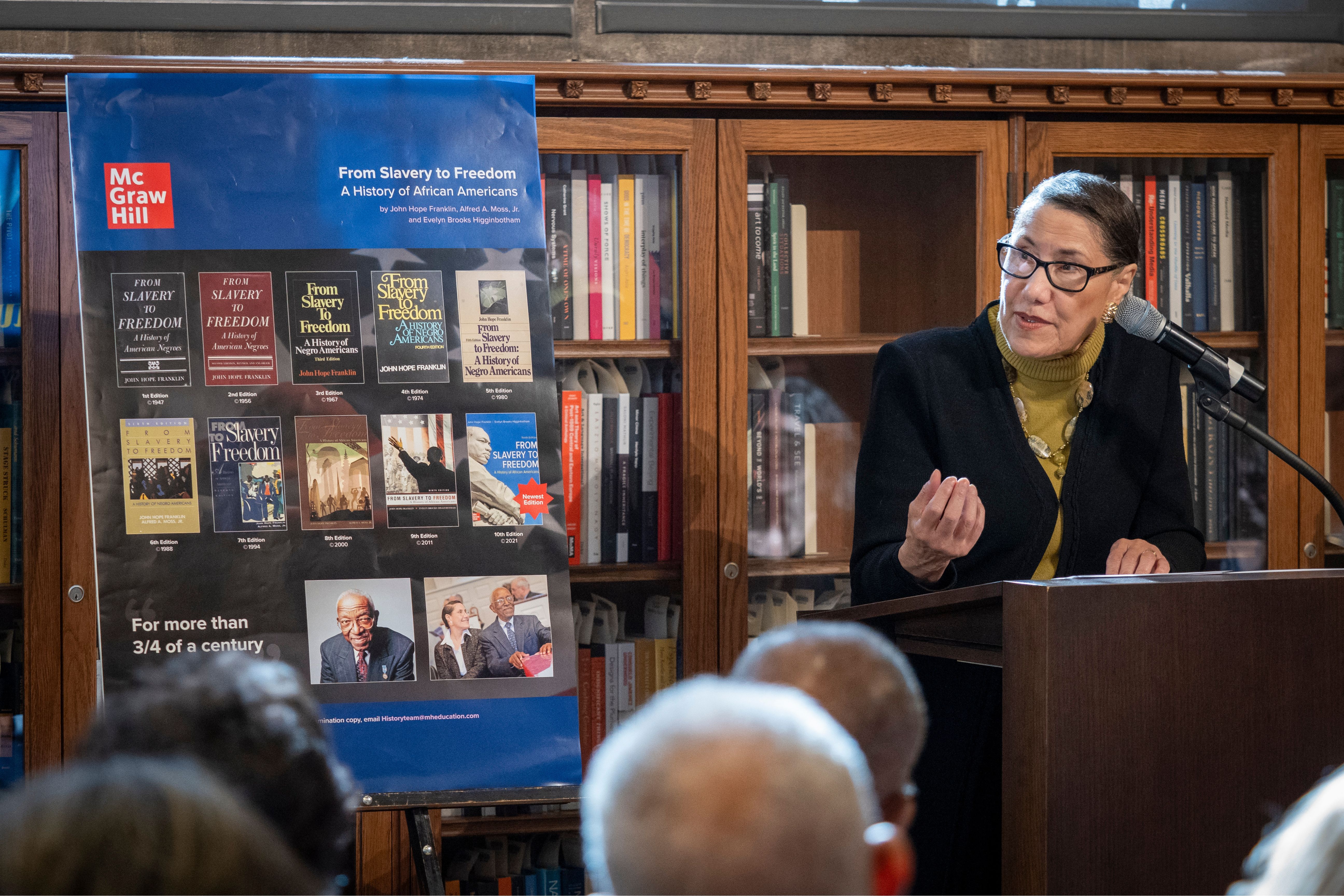 A woman stands at a podium emphasizing a point. She stands next to a poster showing the ten different covers of the editions of John Hope Franklin's book,  "From Slavery to Freedom"