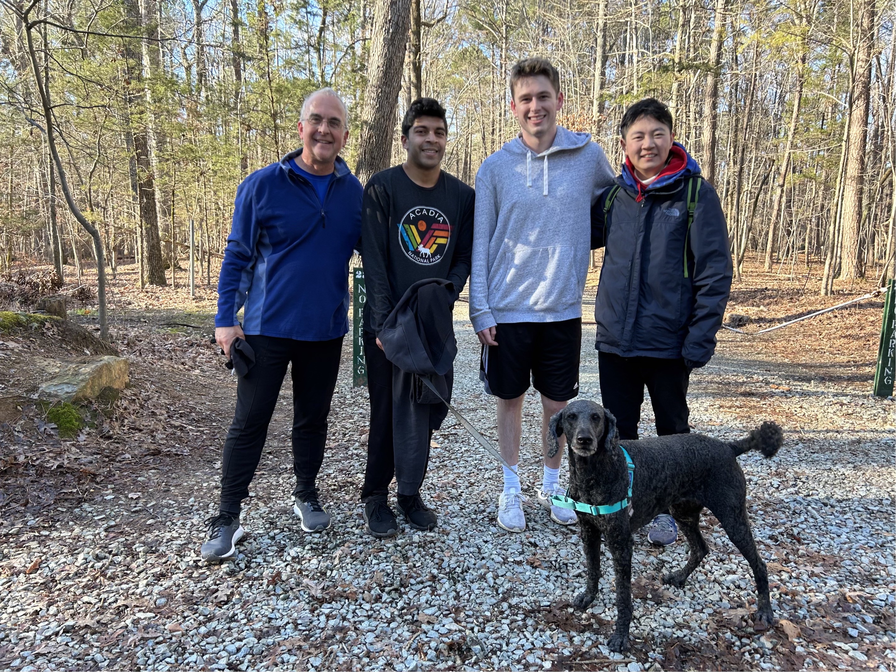 Four men in running clothes stand on a path in the forest. One of them has a black dog on a leash. Len White is at left.