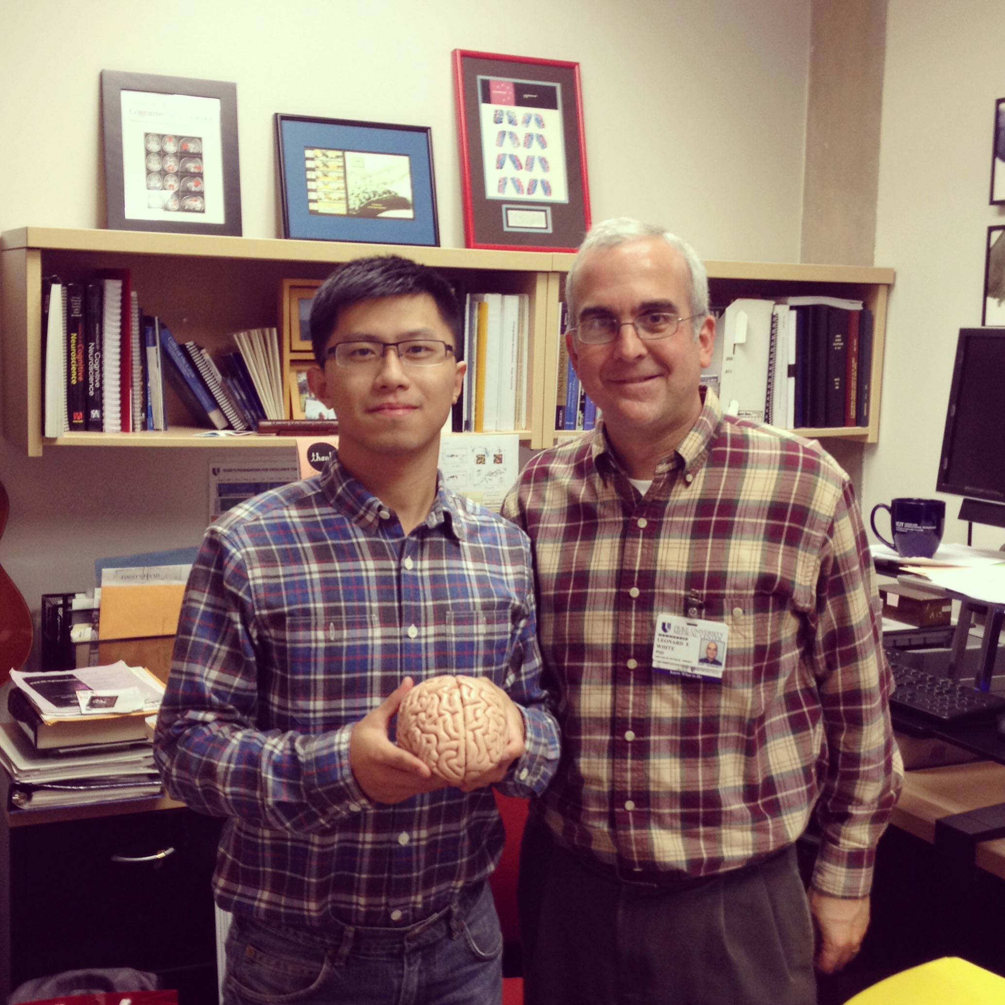 Two men in plaid shirts stand next to each other in an office. The man on the left is holding a human brain. 