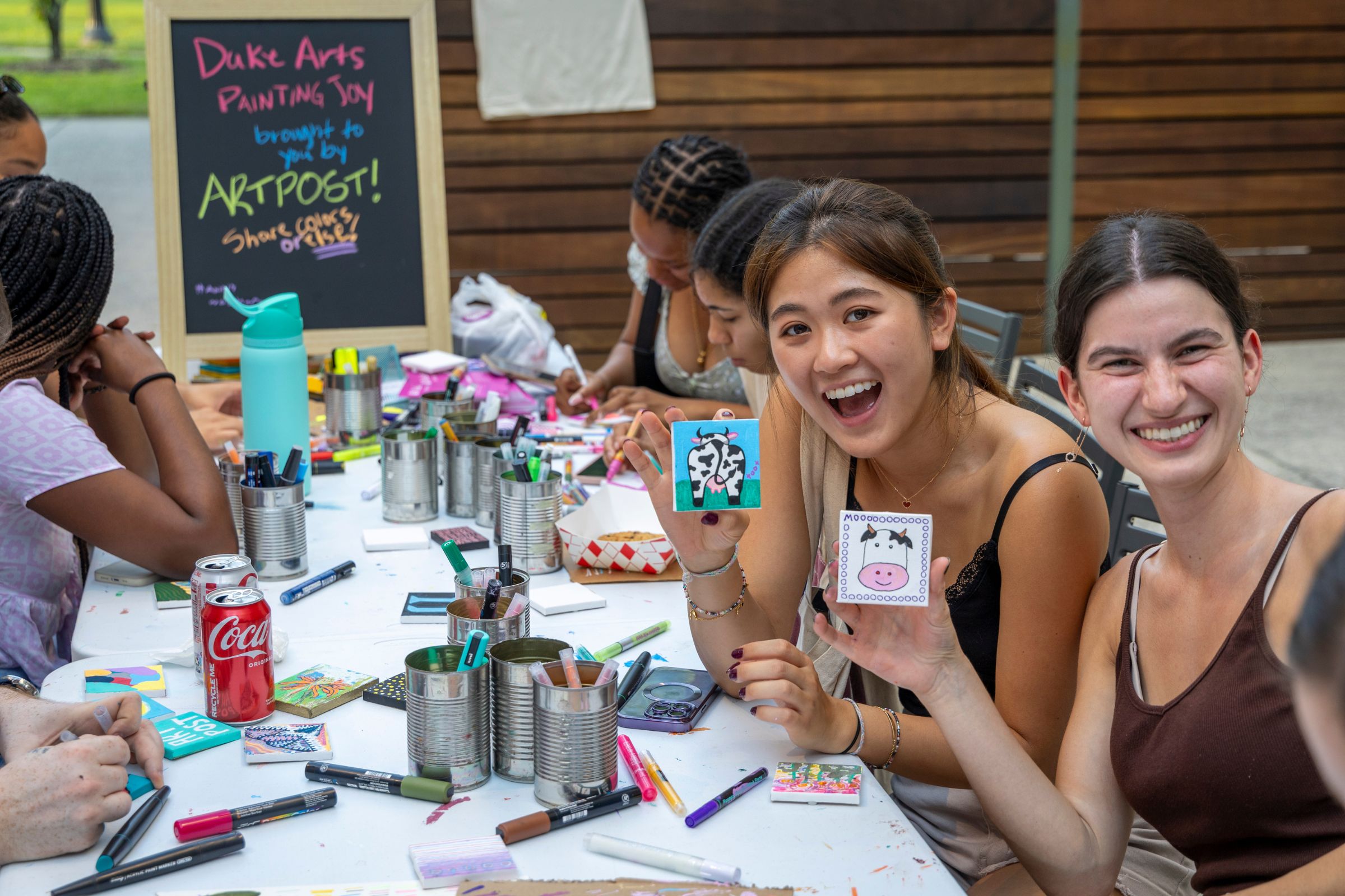 Two young women, at a table covered with arts supplies, smile and show their arts tiles on which they have just painted funny cows. One tile has a smiling cow face and the other the cow's behind, tail and udder.
