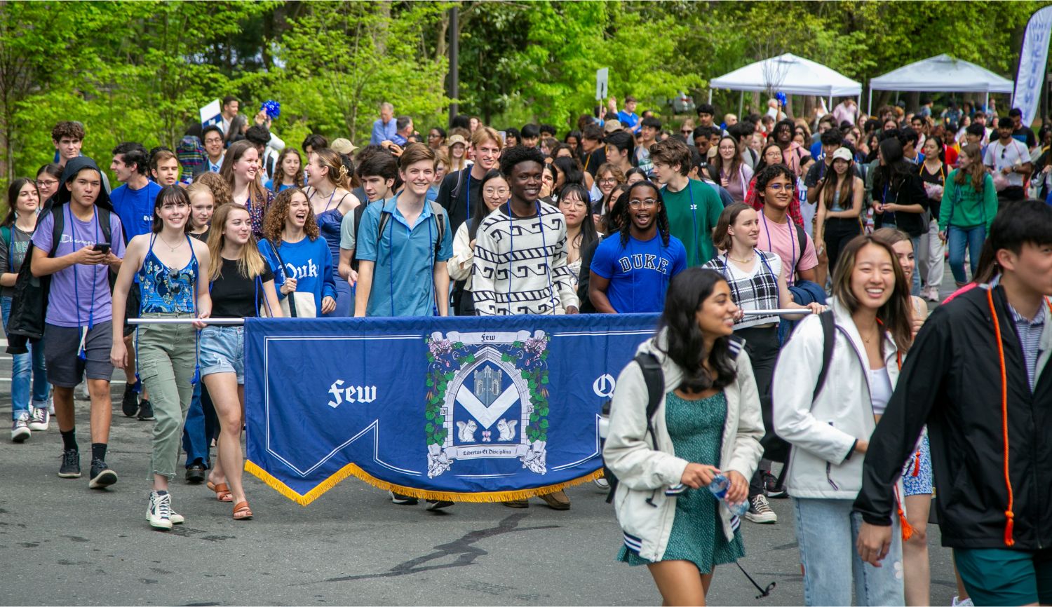 Students walk up Chapel drive laughing and smiling. Students carry a long dark blue banner with the shield for Few Quad.
