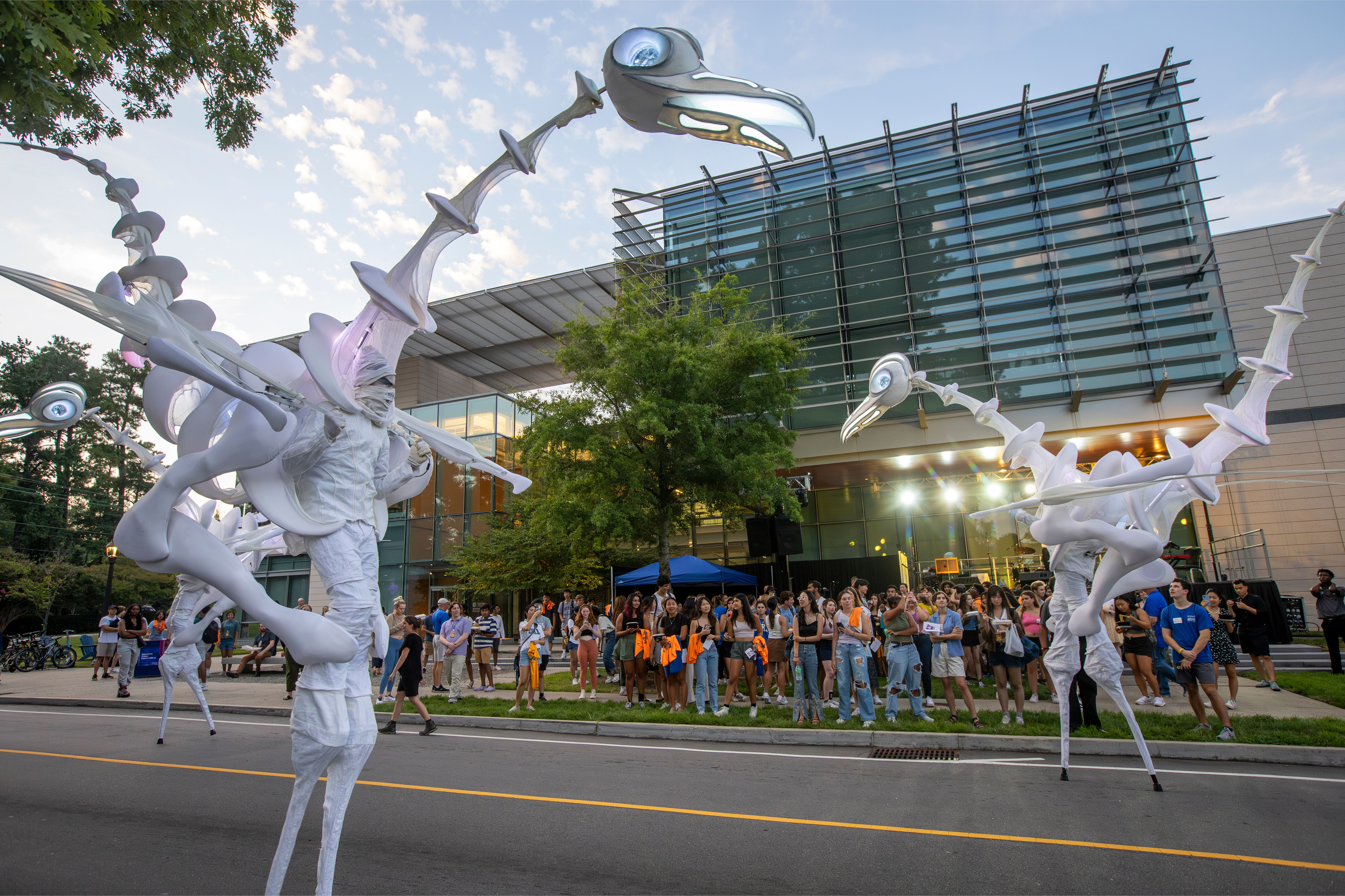 Three, white "birdmen" puppets walk in a circle on the street in front of a crowd of students at the Rubenstein Arts Center