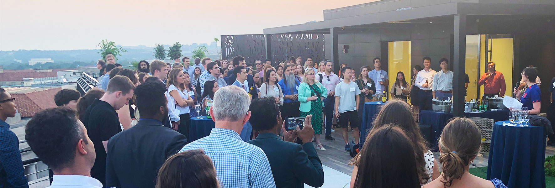 A large gathering of people stand outside on a rooftop patio watching a speaker.