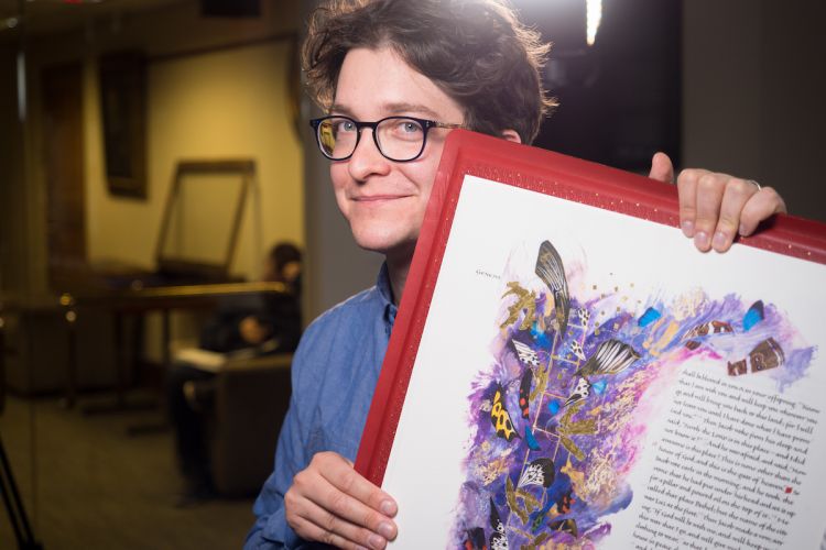 A man with glasses holds a large book with colorful images and calligraphy.