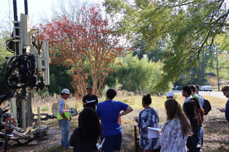 Students stand near large drill and engineer