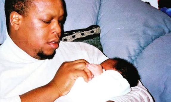 Mark Anthony Neal sits on comfortable furniture and holds an infant. Both are wearing white.