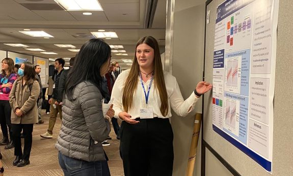 Alyssa Guthrie in black pants and a white blouse presenting her research poster to a peer.