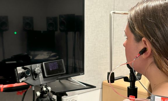 Co-first author Stephanie Lovich shows what participants in their research study did and stares at a green dot on a TV while her chin rests in a holder to keep her head still while tracking the dot w/ her eyes