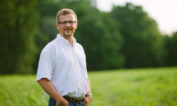 Norman Wirzba, hands in the pockets of his bluejeans, stands in a green field. 