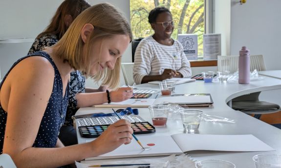 A woman paints a watercolor while two other women in the background look on