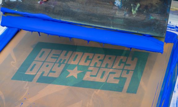 A hand holds a paint scraper over a screen print screen with blue paint dripping. the screen says "Democracy Day 2024"