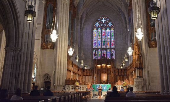 The interior of Duke Chapel shows a stained glass windows behind an altar beneath a Gothic arch
