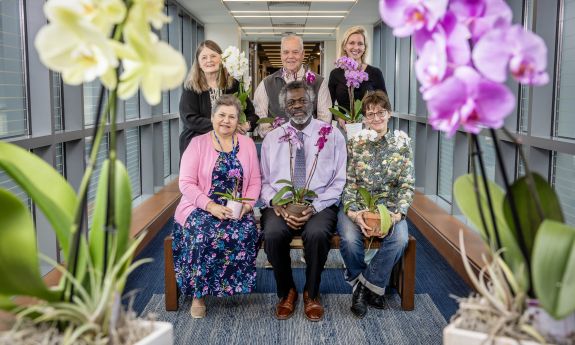 Six staff and faculty members pose in a group holding orchid plants, framed by two orchids on either side of them