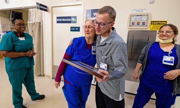 Three women in scrubs and a man in a button-down shirt stand in a hallway, as the man hugs one of the women