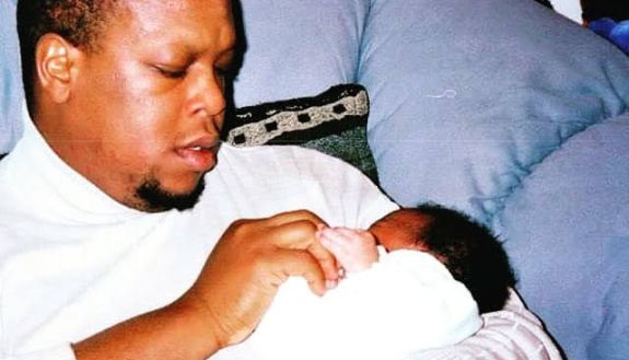 Mark Anthony Neal sits on comfortable furniture and holds an infant. Both are wearing white.