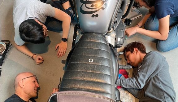 Overhead view of three students and instructor sitting or lying next to a motorcycle they are working on.