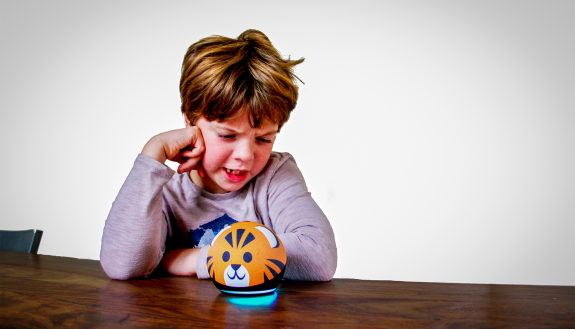 An 8-year-old boy at a table, his head resting on his fist, appears to be having angry words with a kid-friendly Alexa device that looks like a cat's head.