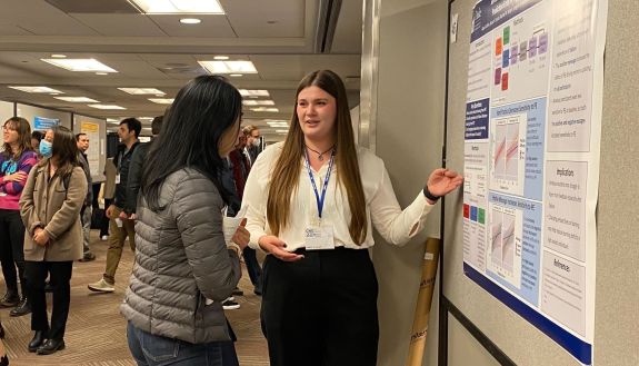 Alyssa Guthrie in black pants and a white blouse presenting her research poster to a peer.