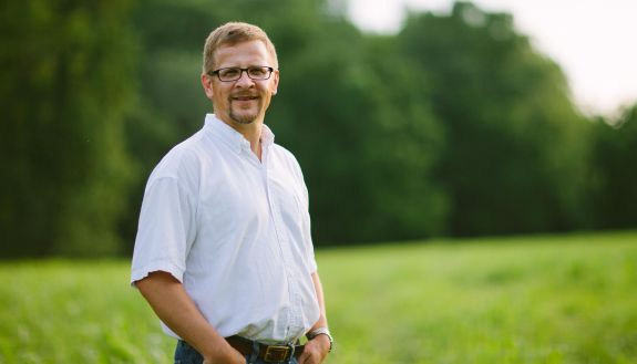 Norman Wirzba, hands in the pockets of his bluejeans, stands in a green field. 