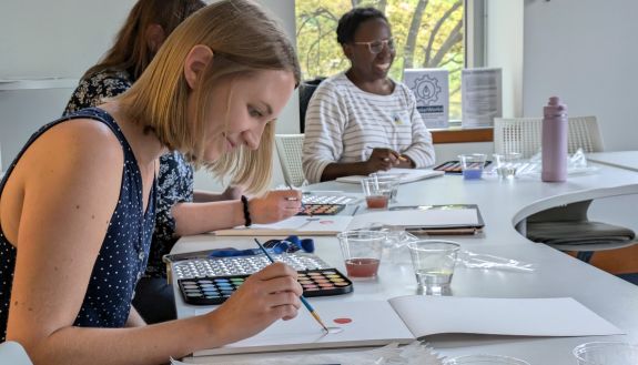 A woman paints a watercolor while two other women in the background look on