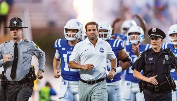 Duke head football coach Manny Diaz runs onto the field, flanked by police officers and followed by players in uniform 