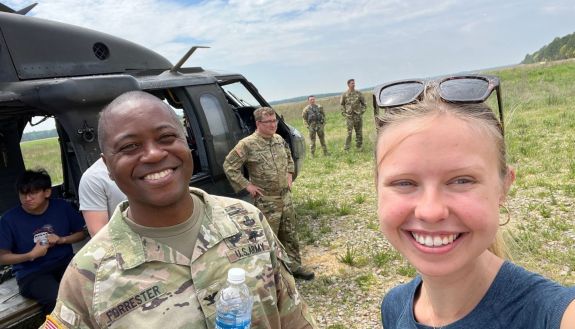 people in military uniform in a field by a helicopter, with student on the right 