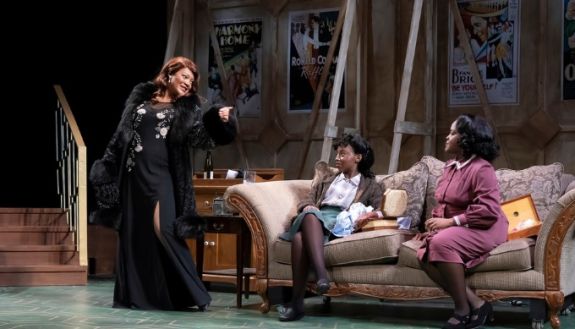 Actors on a stage, two are on a couch and one is standing