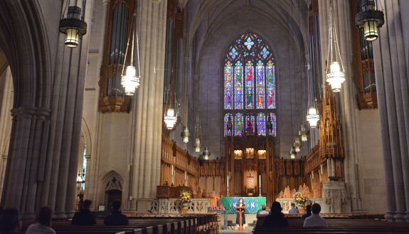 The interior of Duke Chapel shows a stained glass windows behind an altar beneath a Gothic arch