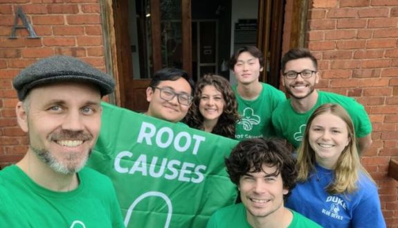 A photo of people in green shirts holding a sign that says "Root Causes"