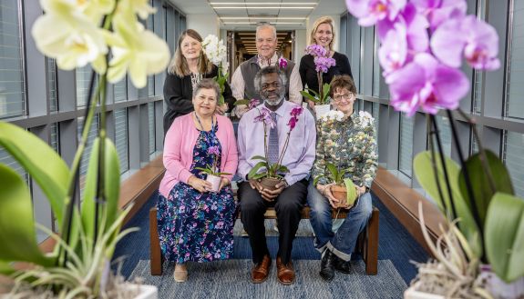 Six staff and faculty members pose in a group holding orchid plants, framed by two orchids on either side of them